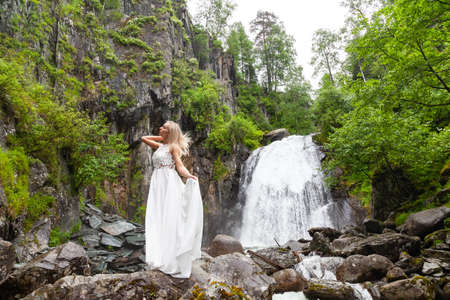 A young blonde girl standing in a half-turn, straightening hair in a boudoir dress in the mountains against a waterfall and stones like a ballerina on a warm summer day in the Altai green treeの写真素材