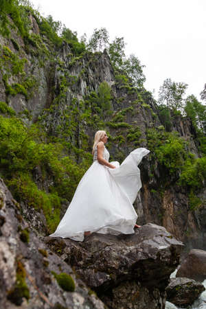 A young blonde girl in an elegant pose throws up hem of a boudoir dress in the mountains against a waterfall and stones raising her hands up like a ballerina on a warm summer day in Altai green treesの写真素材