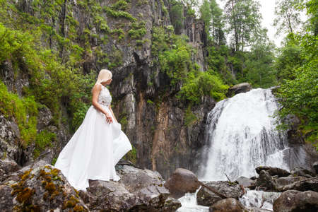 A young blonde girl in an elegant pose hold in arms hem of a boudoir dress in the mountains against a waterfall and stones looking back like a ballerina on a summer day in Altai green treesの写真素材