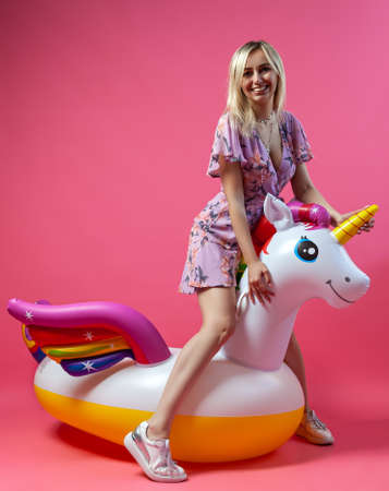 A beautiful blonde girl in a sexy sundress with slim legs in white sneakers stands on an inflatable multi-colored unicorn on a pink isolated background. Summer vacation at the beach and travel.の写真素材