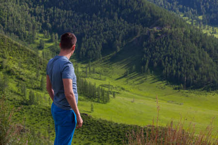 Guy in a green t-shirt is standing  on the edge of the cliff in rear of him is a colorful view of...