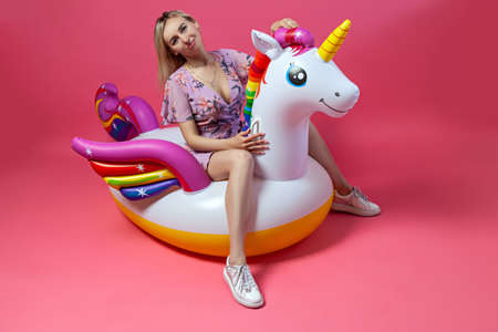 A beautiful blonde girl in a sexy sundress with slim legs in white sneakers sits on an inflatable multi-colored unicorn straightens hair on a pink isolated background. Summer vacation at the beachの写真素材