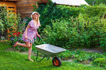 Young beautiful girl blonde in a dress and hat, having fun in the garden holding in her hands a green cart on the lawn with grass on the bushes background.の写真素材
