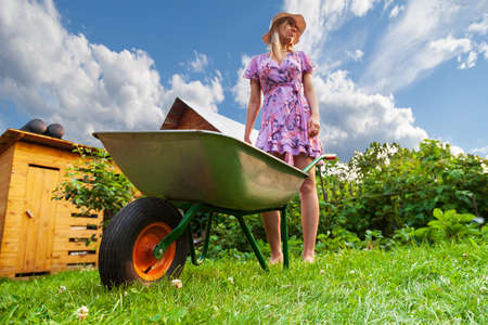 Young beautiful girl blonde in a dress and hat, having fun in the garden holding in her hands a green cart and looking to the side on the lawn with grass on the bushes background.の写真素材