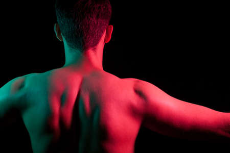 Close-up of nape, back and shoulders with strained muscles and hands apart of an short hair head of a man with stubble, naked without clothes, illuminated in red and green on black isolated backgroundの写真素材