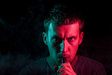 A young brunette man holds in his hand and drags on an electronic cigarette with a vape while...