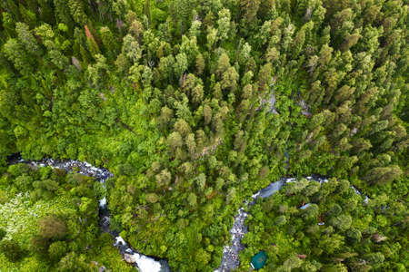 A large waterfall in the back of the Altai Mountains near a steep cliff with green trees, house and green roof. Rest and loneliness while traveling to deserted places. Aerial viewの写真素材