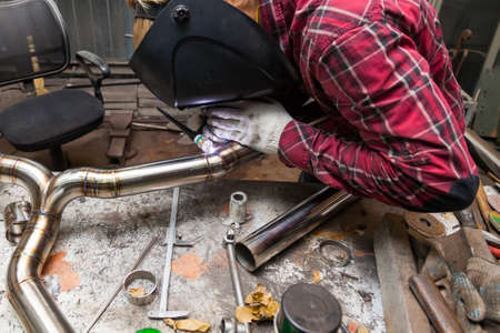 Young guy welder in a checkered red shirt welds a stainless steel pipe using agronomic welding to...