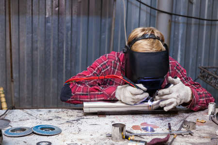 Young guy welder in a checkered red shirt welds a stainless steel pipe for car exhaust using agronomic welding to protect his eyes with a mask in an iron workshop. Modern welding methods.の写真素材