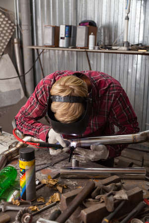 Young guy welder in a checkered red shirt welds a stainless steel pipe using agronomic welding to...