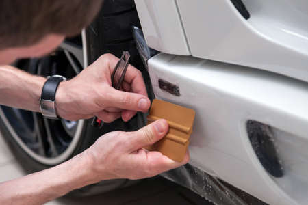 Retrofitting the car with a solid transparent protective film, the master smooths the surface by squeezing air bubbles with a scraper, protecting the vehicles from scratches and chips in the workshopの写真素材