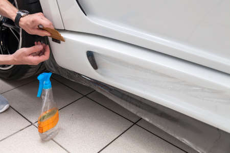 Retrofitting the car with a solid transparent protective film, the master smooths the surface by squeezing air bubbles with a scraper, protecting the vehicles from scratches and chips in the workshopの写真素材