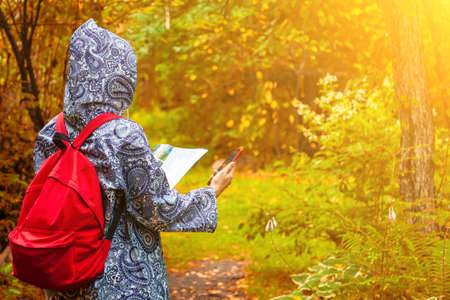 A view from the back of a young blonde girl in a blue raincoat with a red backpack on her back, a phone in her hand walks along a path and looking to the map in a park or forest. Travelling in wild.の写真素材