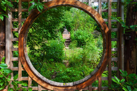 View through a round wooden window of a Buddha statue sitting in a lotus position for worship and meditation of Buddhists in a flowering green garden with green trees and leaves.の写真素材