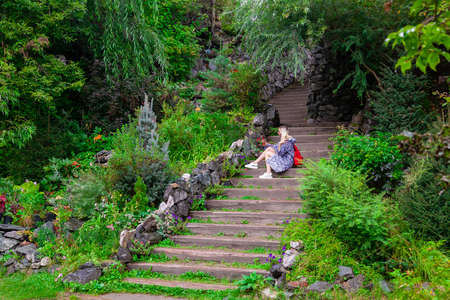 Young beautiful blonde girl in a blue raincoat sits on a wooden staircase with stone walls in a...