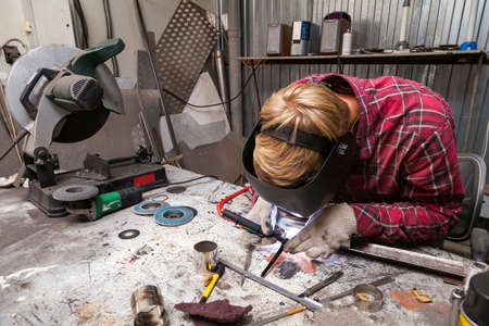 Young guy welder in a checkered red shirt welds a stainless steel pipe using agronomic welding to...
