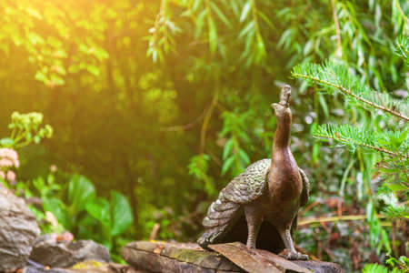 A stone statue in the form of a sacred animal peacock in India and Hinduism sitting on a stone with his head turned in the opposite direction against a background of green leaves.の写真素材