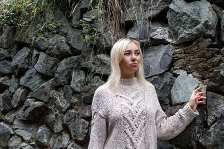 Pastel color image of a young blonde girl in a gray knitted sweater on a background of a stone wall in the fall. Fashionable clothes and style in the cold season.の写真素材