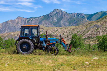 An old blue tractor excavator stands in a field with green and yellow grass against a background of mountains and blue sky during harvesting in summer or autumn. Agriculture and farming machinery.の写真素材