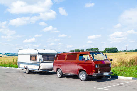 A motor home in the form of a white trailer in a parking lot near the road, fastened to a red car in the back of a minivan while traveling on a summer day against a blue sky. Movable property.の写真素材