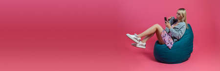 Beautiful blonde girl in a blue jacket and a purple sundress sits on a green bag chair with her lifting legs on a pink isolated background and talking on the phone and looking to a smartphoneの写真素材