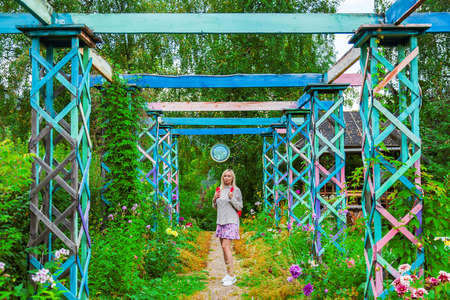 A young blonde girl in a knitted sweater with a red backpack on her back, a phone in her hand walks along a path in wooden colored arch  in park or forest. Travelling in wildの写真素材