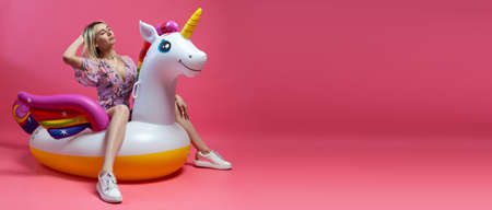 A beautiful blonde girl in a sexy sundress with slim legs in white sneakers sits on an inflatable multi-colored unicorn on a pink isolated background. Summer vacation at the beach and travel.の写真素材