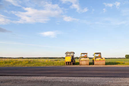 Three large yellow asphalt compactor standing on the side of the track during highway road repair and laying black new asphalt against a landscape with a blue sky and clouds in the summer.の写真素材