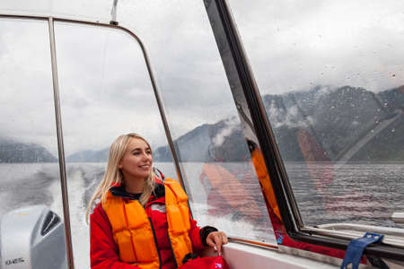 Beautiful young blonde woman in an orange life jacket sits in a motor boat during an excursion and...