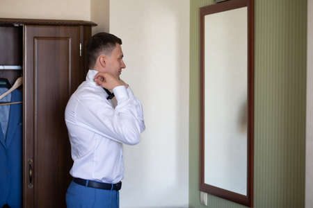 A young man husband is going to a wedding ceremony in a luxury hotel wearing a blue suit and adjusting his bow tie while waiting for the bride. Love and lovestory during the groom's gatherings.の写真素材