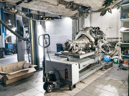 A used car raised on a lift for repair and under it a detached engine and gearbox suspended on a...