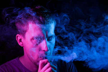 Close up portrait of the face of an adult serious man exhales blue toxic smoke while smoking e-cigarette and vape illuminated with pink colored light on a black background. Harm to health.の写真素材