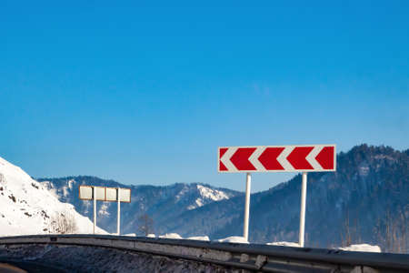 Road sign warning of a sharp and dangerous turn to the left with red arrows on a white background with mountains, snow in winter.の写真素材