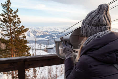 A young girl in a gray knitted hat looks through stationary binoculars on an observation deck in the...