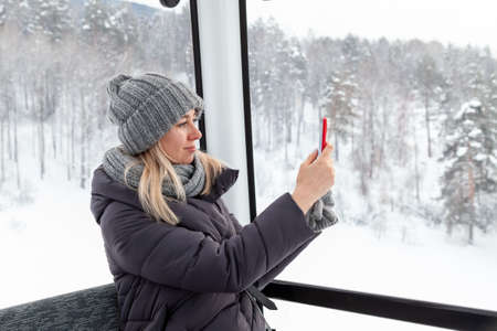 A young beautiful blonde girl in winter clothes and a gray knitted hat sits in the gondola lift...