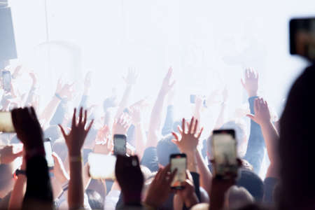 Audience with hands raised at a music festival with colored lights streaming down from above the stage. Rock or rap concert with many people.の写真素材