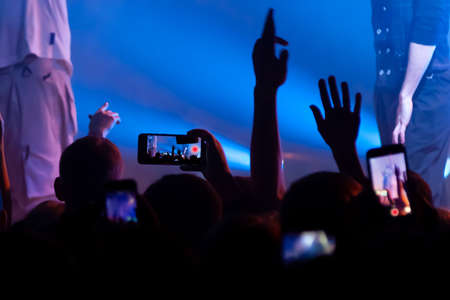 Hand with a smartphone records live music festival, Taking photo of concert stage. Party at nightclub with crowd of people raising hands with phones.の写真素材