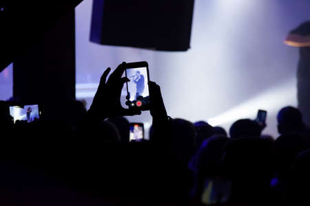 Hand with a smartphone records live music festival, Taking photo of concert stage. Party at nightclub with crowd of people raising hands with phones.の写真素材