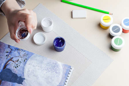 A female hand holds a lid from a jar on which paint is mixed against the background of a cat on a tree, the night sky and the moon next to work tools. Quarantine craze.の写真素材