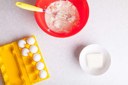 On the kitchen table is a red bowl with kneading dough from cottage cheese and with a whisk, a white...