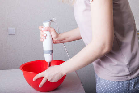 Woman in the kitchen mixes ingredients for panettone dough in a red bowl holding a submersible electric blender in hands. The daily routine of the wife and mother is cooking for the family.の写真素材