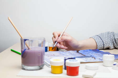 Female hand holds a brush and draws picture at table in middle of jars with watercolor paints and next to it is a glass for rinsing and cleaning the brush from color on a white background in studio.の写真素材