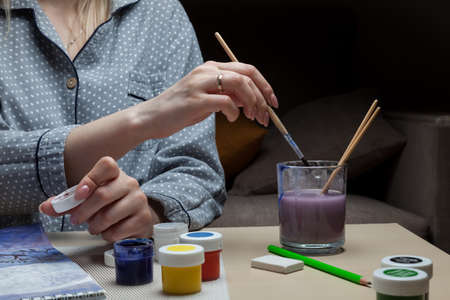A lesson in long cozy winter evenings - a girl sits at a table and dips a brush in a glass to clean it from pigment, in her other hand she holds a lid of watercolors, next to are cans of paint.の写真素材