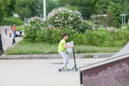 Novosibirsk, Russia - 06.06.2020: A teenager boy on a scooter picks up speed to ride a ramp and jump to the top in the park against a background of green trees.の写真素材