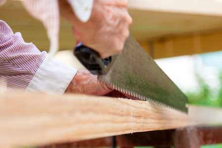 A metal hand saw with a black plastic handle with sharp teeth in the hands of a carpenter when sawing wooden beams on a summer day during construction on a summer cottage.の写真素材