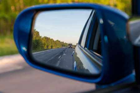 Rear view in the side mirror of a blue sedan with the reflection of a gray truck on an asphalt road on a summer day with green trees on the sides of the highway.の写真素材
