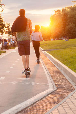Couple of Teenagers ride on electric scooters on Mikhailovskaya Embankment, many people walk in the park on weekends at sunset on a warm summer day.の写真素材