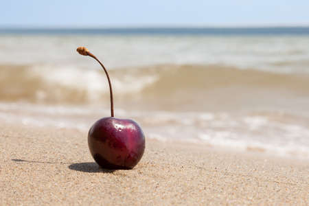 One berry of ripe red cherries on the beach by the sea on the sand, a romantic seductive dessert on vacation against the backdrop of waves on a sunny day.の写真素材