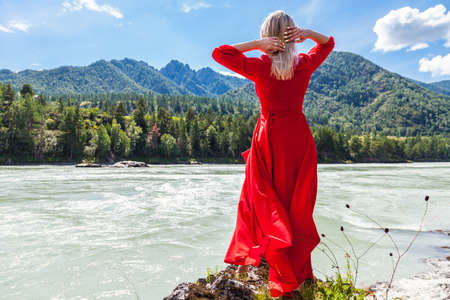 Back view of a blonde woman in a long red dress with her hands thrown back behind her head against the background of the river, mountains covered with green forest, and the blue sky in Altai.の写真素材