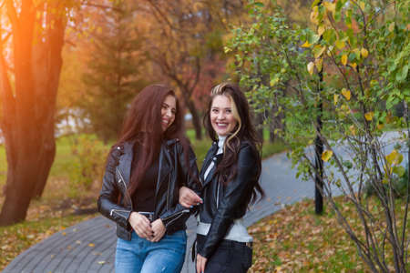 Two girlfriends, students laugh and stand on the path in jackets and jeans while walking in the park...